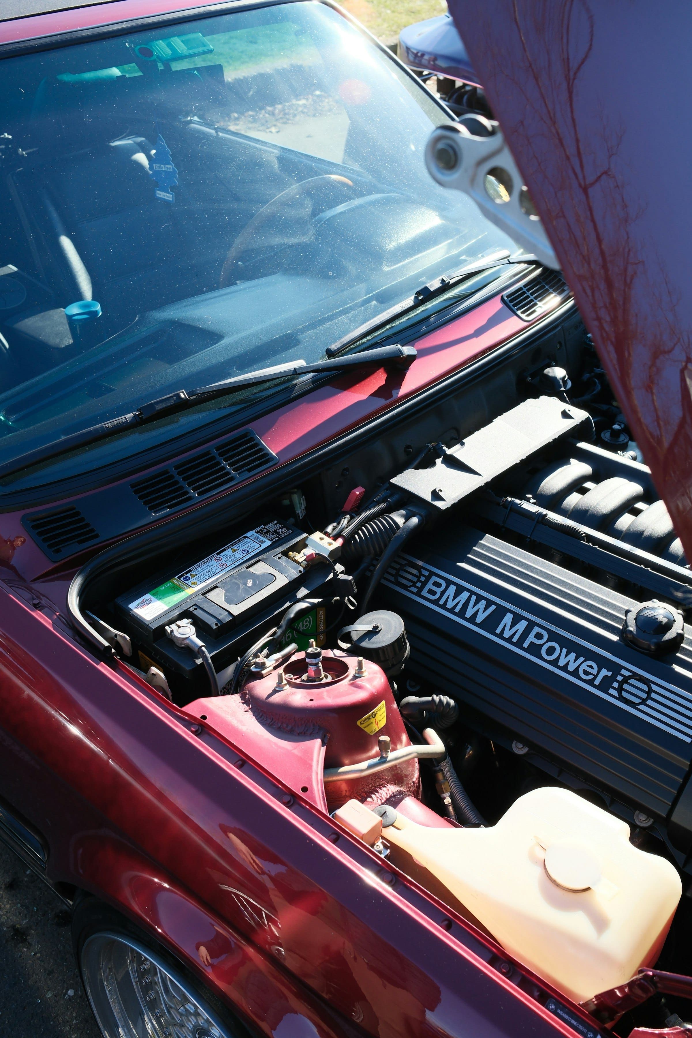 The open bonnet of a red car in bright sunlight, showing the engine bay with the battery and air intake
