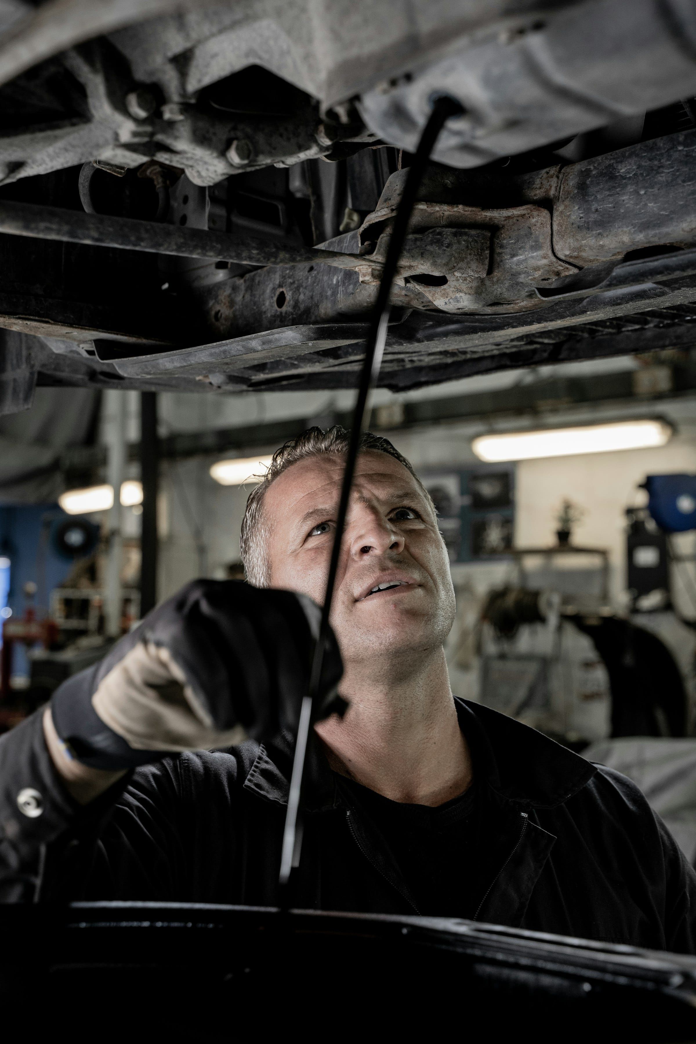 A mechanic in gloves looks up at the underside of a car in a dimly lit garage